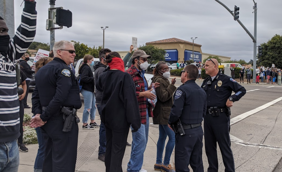 police confronting a few protesters on a street corner, with other protestors on the opposition corner