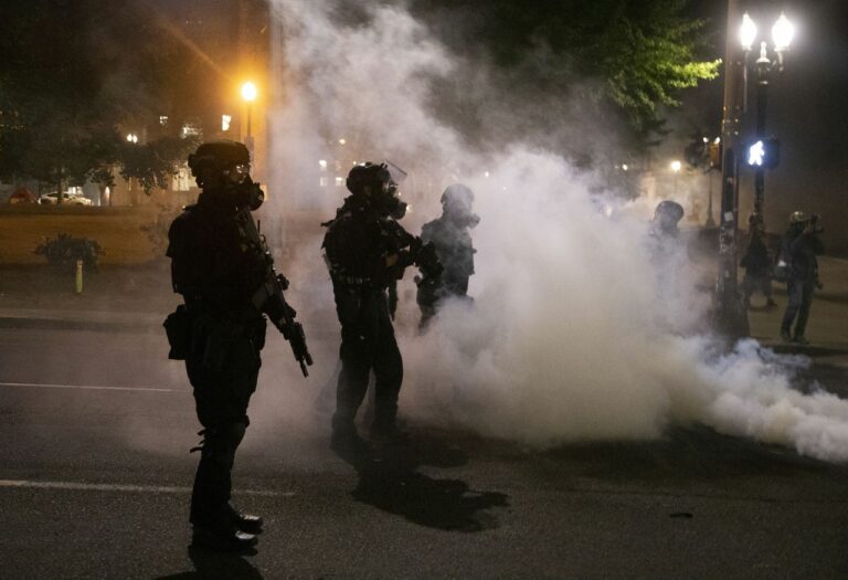 Police officers stand on the street in downtown Portland, Oregon, amid a cloud of gas rising from the ground to the right, outside the frame