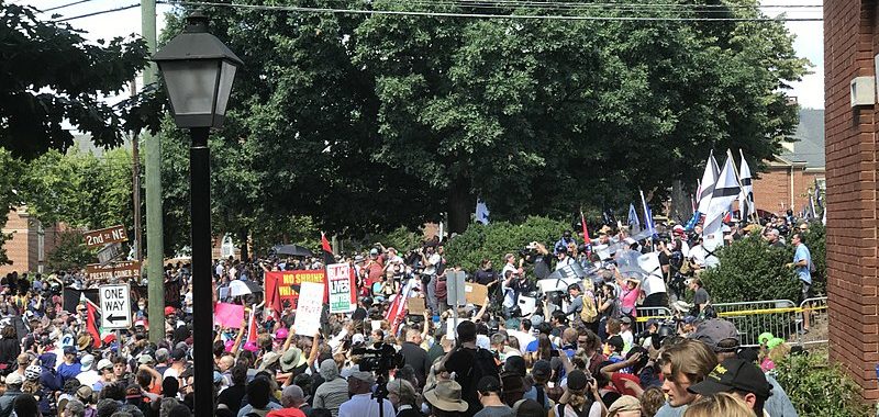 A large crowd of anti-racist protestors face off against white supremacists on a tree-lined street. Only the tops of most people's heads are visible. there are two neon hats in the crowd. They are National Lawyers Guild legal observers.