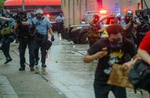 A group of police officers in blue shirts approach down a wet street with guns pointed toward protestors who duck their heads and run away from them. There are cop cars with lights flashing in the background.