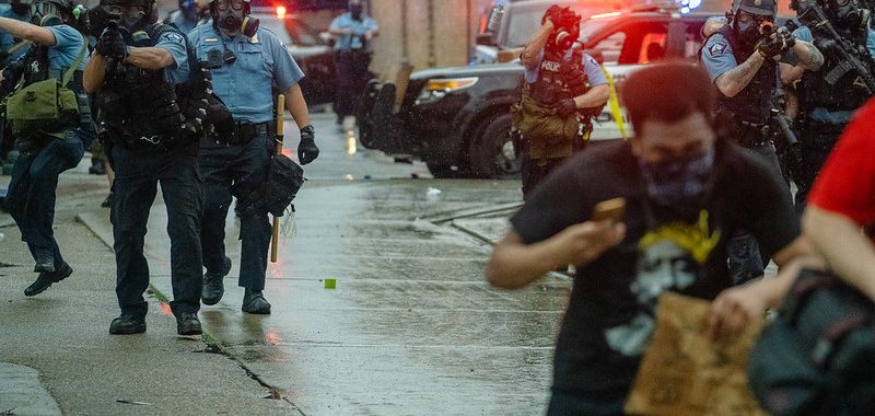 A group of police officers in blue shirts approach down a wet street with guns pointed toward protestors who duck their heads and run away from them. There are cop cars with lights flashing in the background.