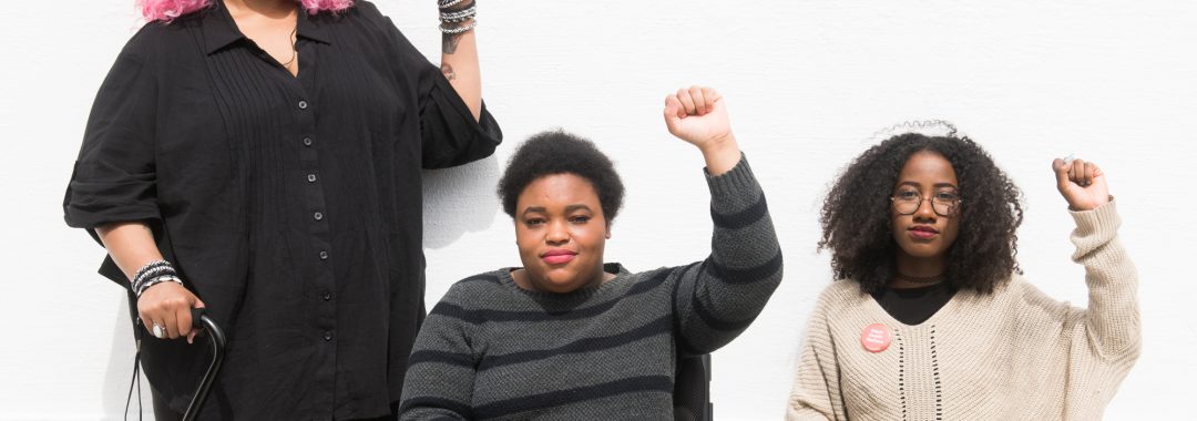 Torso level photo of three Black and disabled folx (a non-binary person holding a cane, a woman in a power wheelchair, and a woman on a folding chair) raising their fists on the sidewalk in front of a white wall. Credit: Disabled and Here