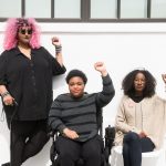 Torso level photo of three Black and disabled folx (a non-binary person holding a cane, a woman in a power wheelchair, and a woman on a folding chair) raising their fists on the sidewalk in front of a white wall. Credit: Disabled and Here
